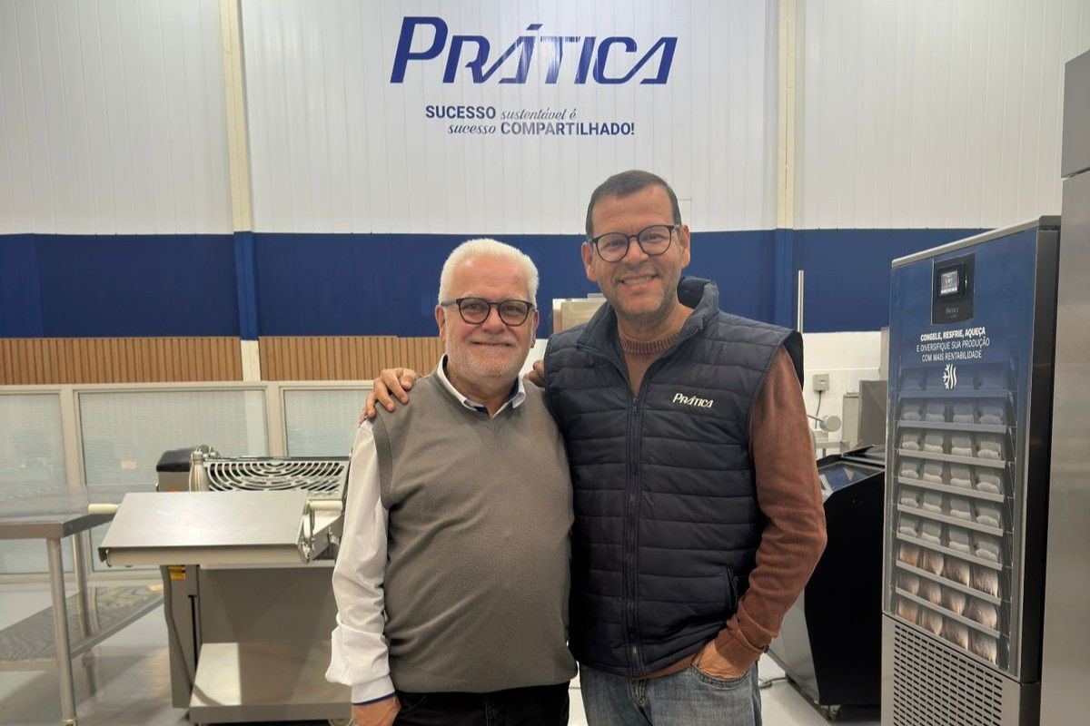 Dois homens posando em frente a uma placa com a inscrição