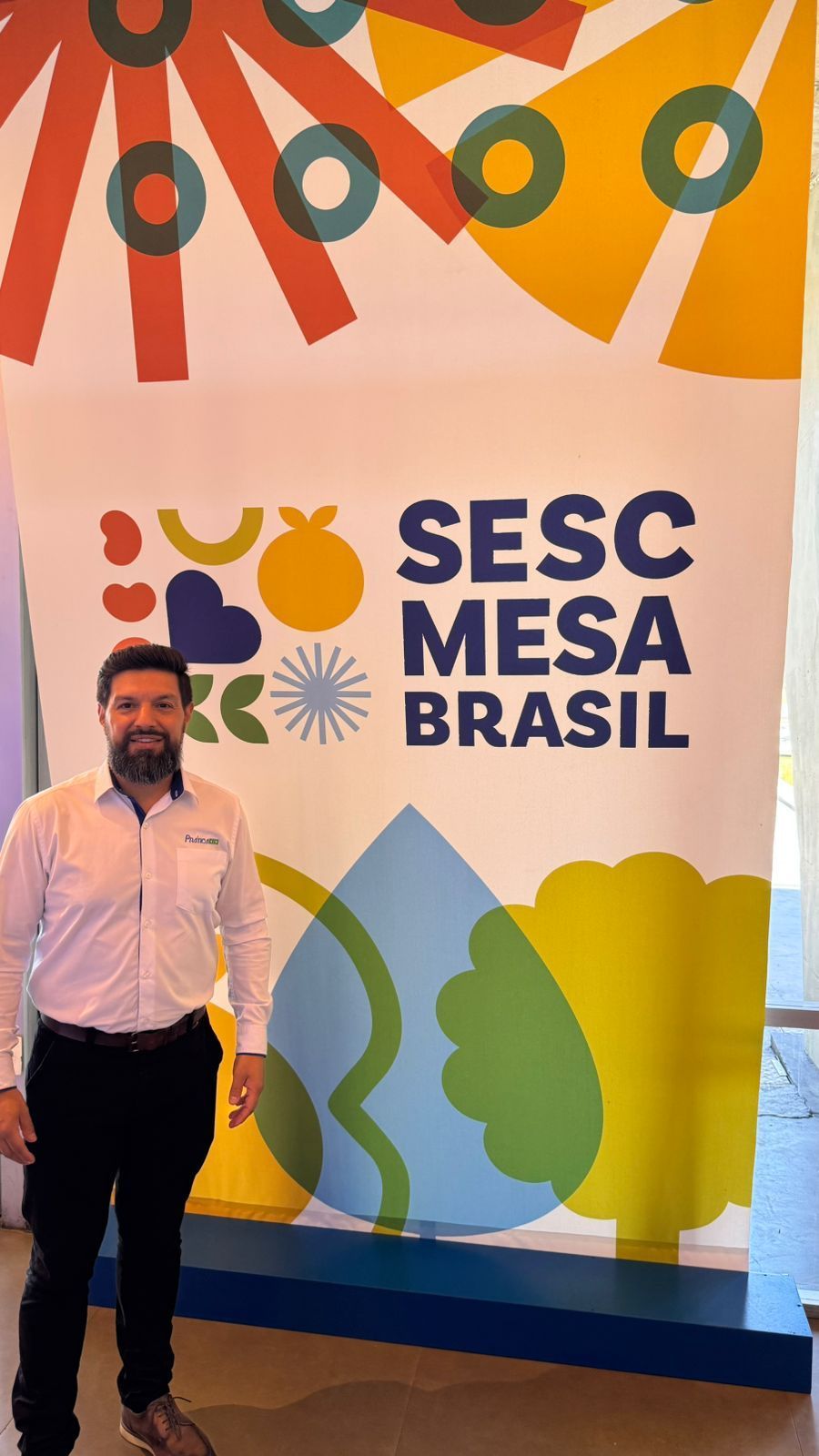 Homem em frente à faixa do SESC Mesa Brasil. Ele sorri e veste camisa branca e calça preta.