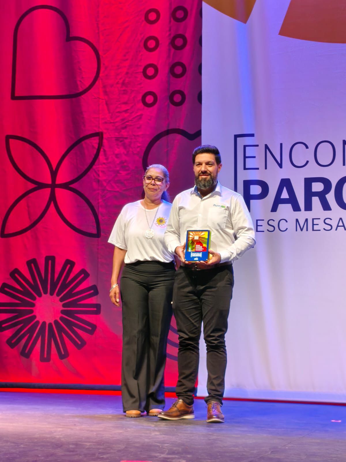 Duas pessoas em um palco durante um evento. Homem segura um prêmio. Fundo vermelho e branco.