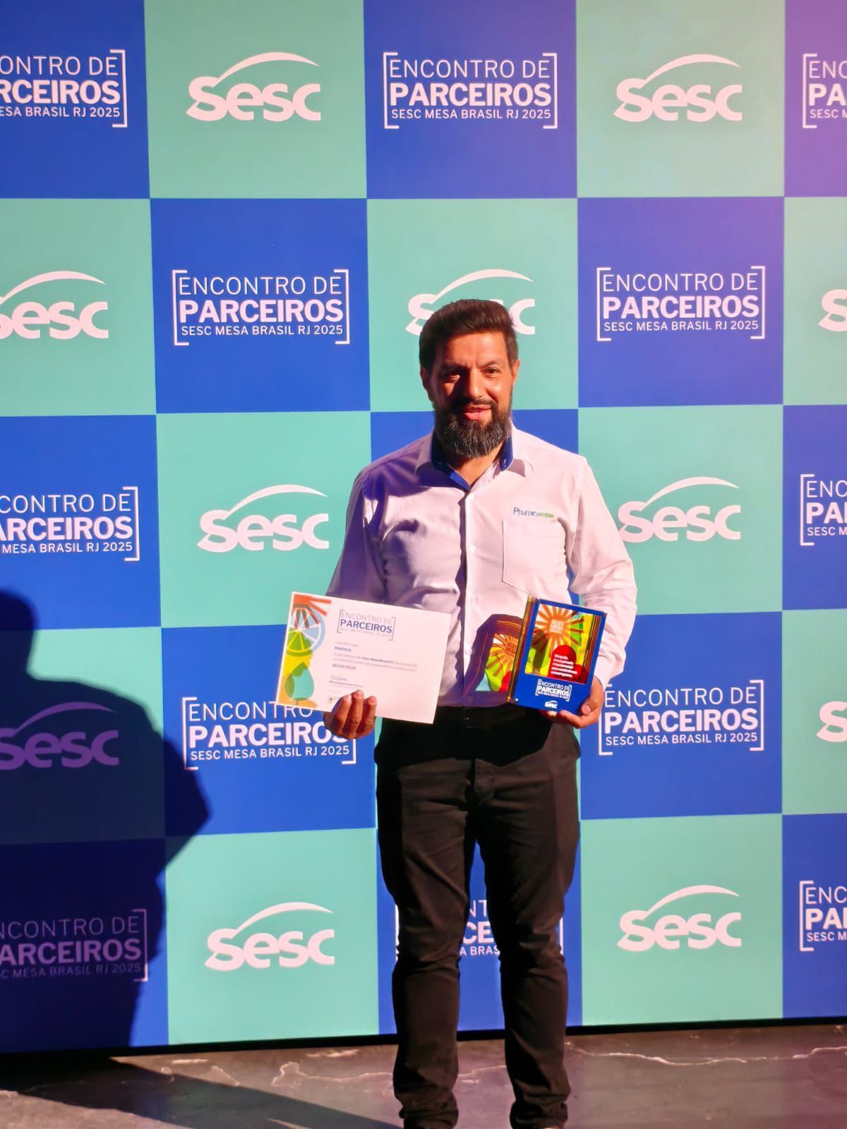 Um homem segurando um certificado e uma caixa, sorrindo, em pé em um palco com faixas azuis do Sesc.