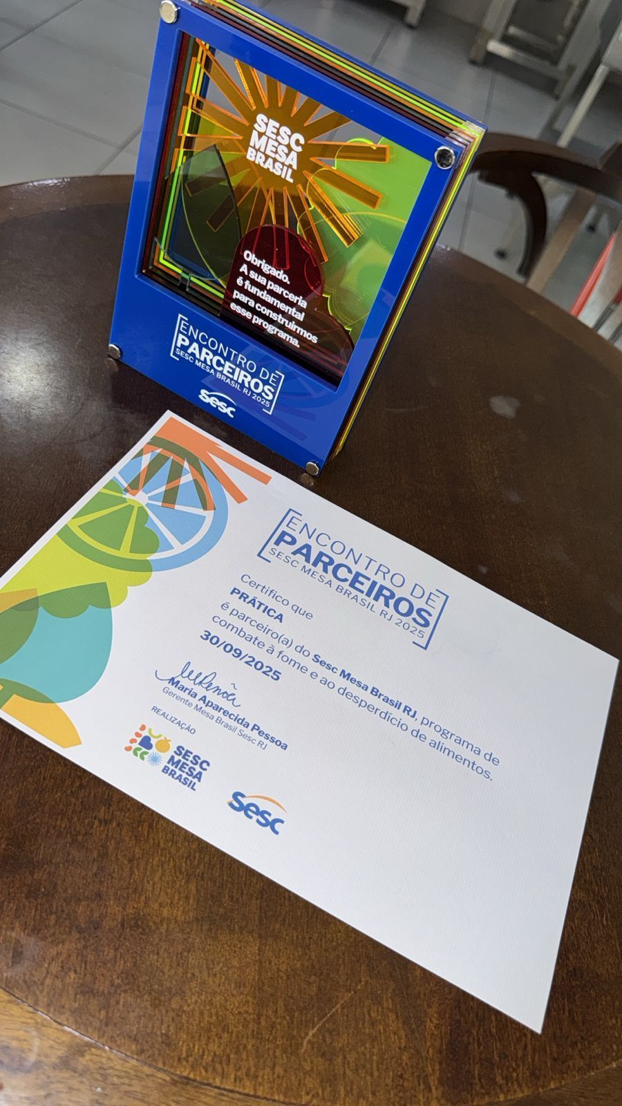 Prêmio e certificado em moldura azul sobre uma mesa de madeira. O certificado tem o logotipo 