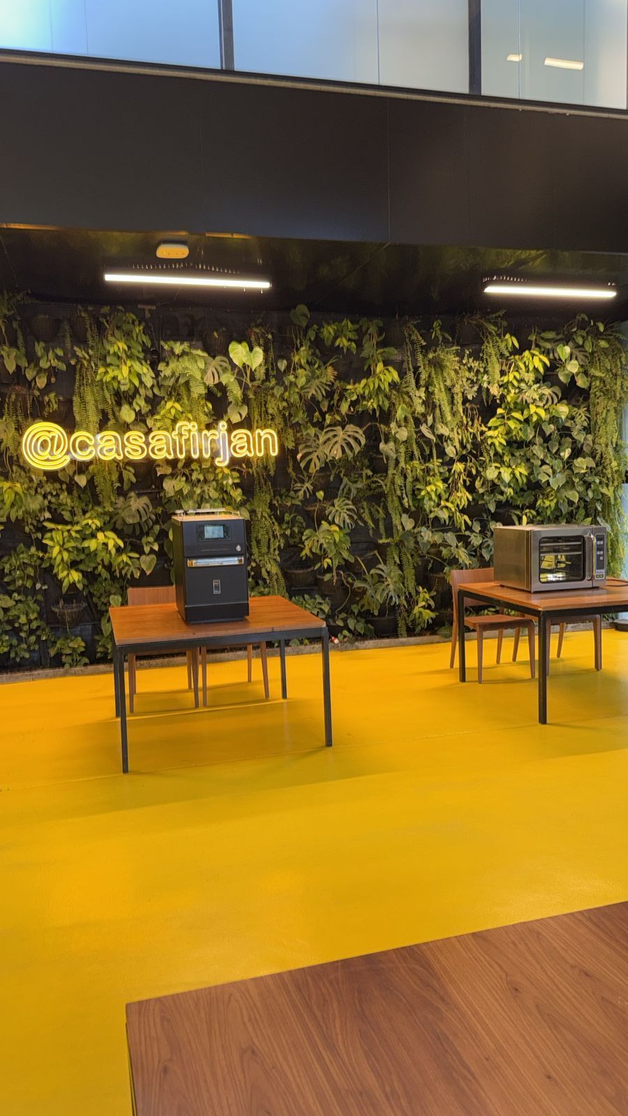 Um interior comercial com piso amarelo, parede de plantas verdes e eletrodomésticos expostos em mesas.