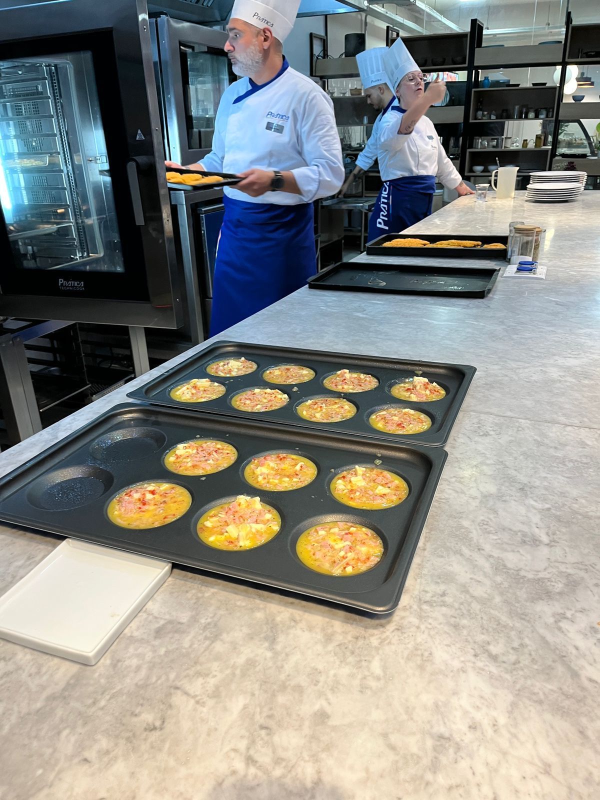 Chefs preparando comida na cozinha, com produtos assados ​​em bandejas no balcão.