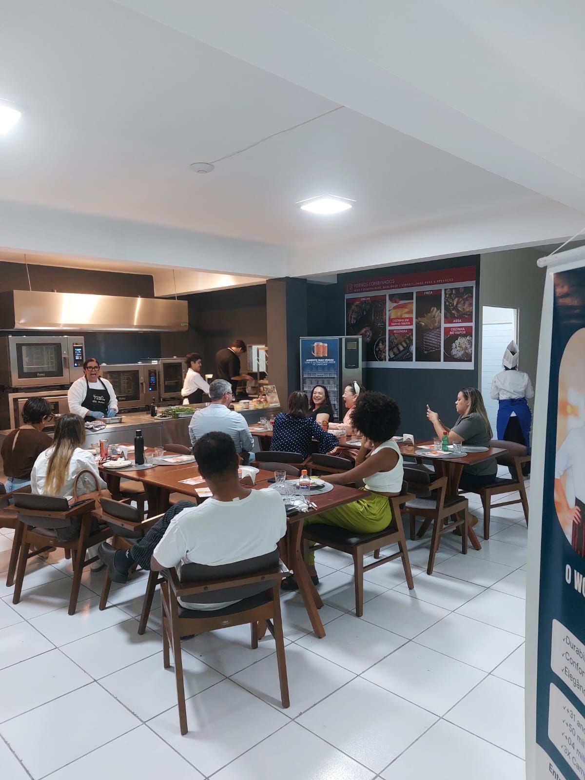 Pessoas sentadas à mesa em uma cozinha, ouvindo um chef demonstrar técnicas de culinária.