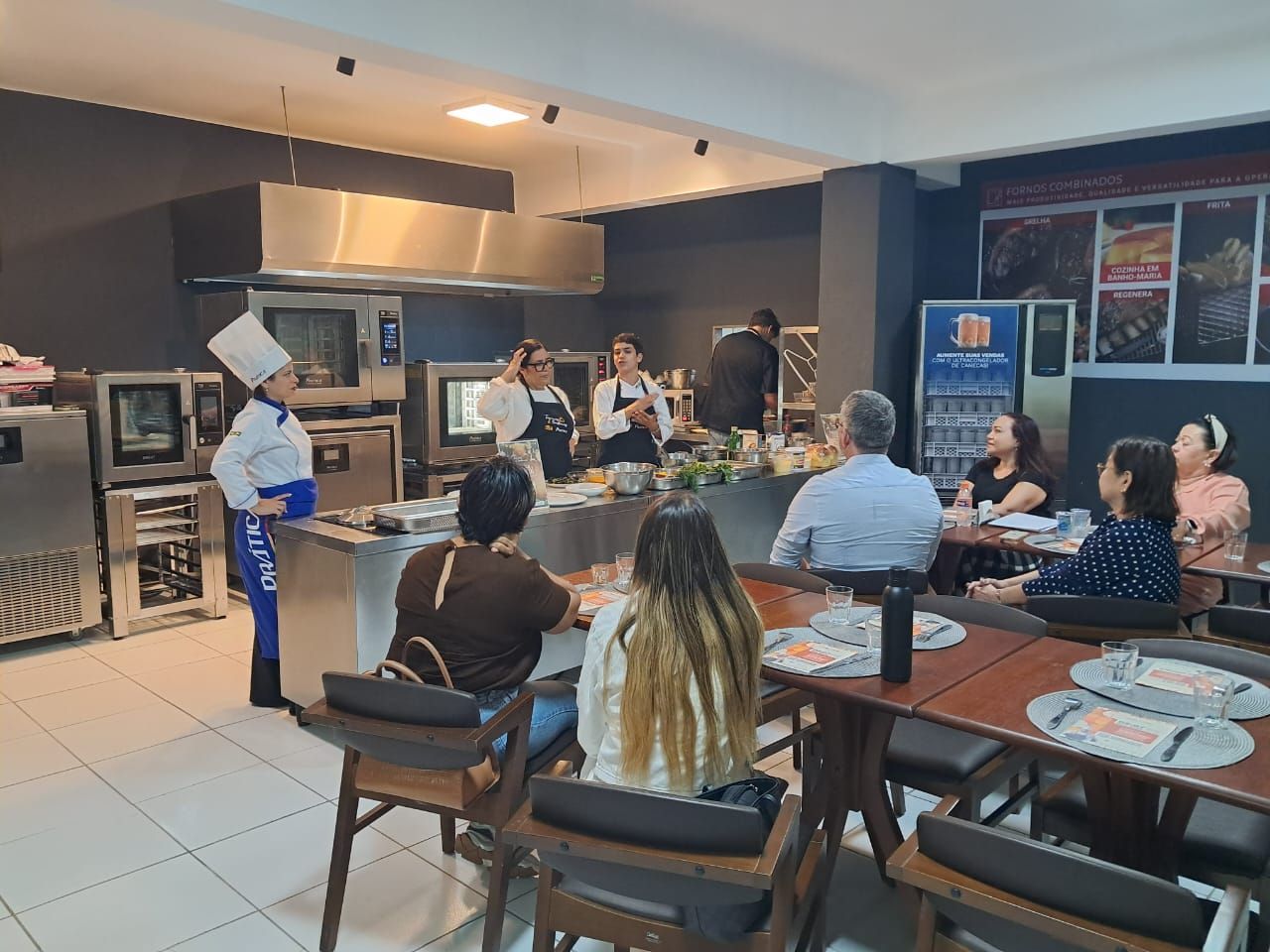 Aula de culinária: Chef demonstra para o público sentado. Cozinha com eletrodomésticos de aço inoxidável.