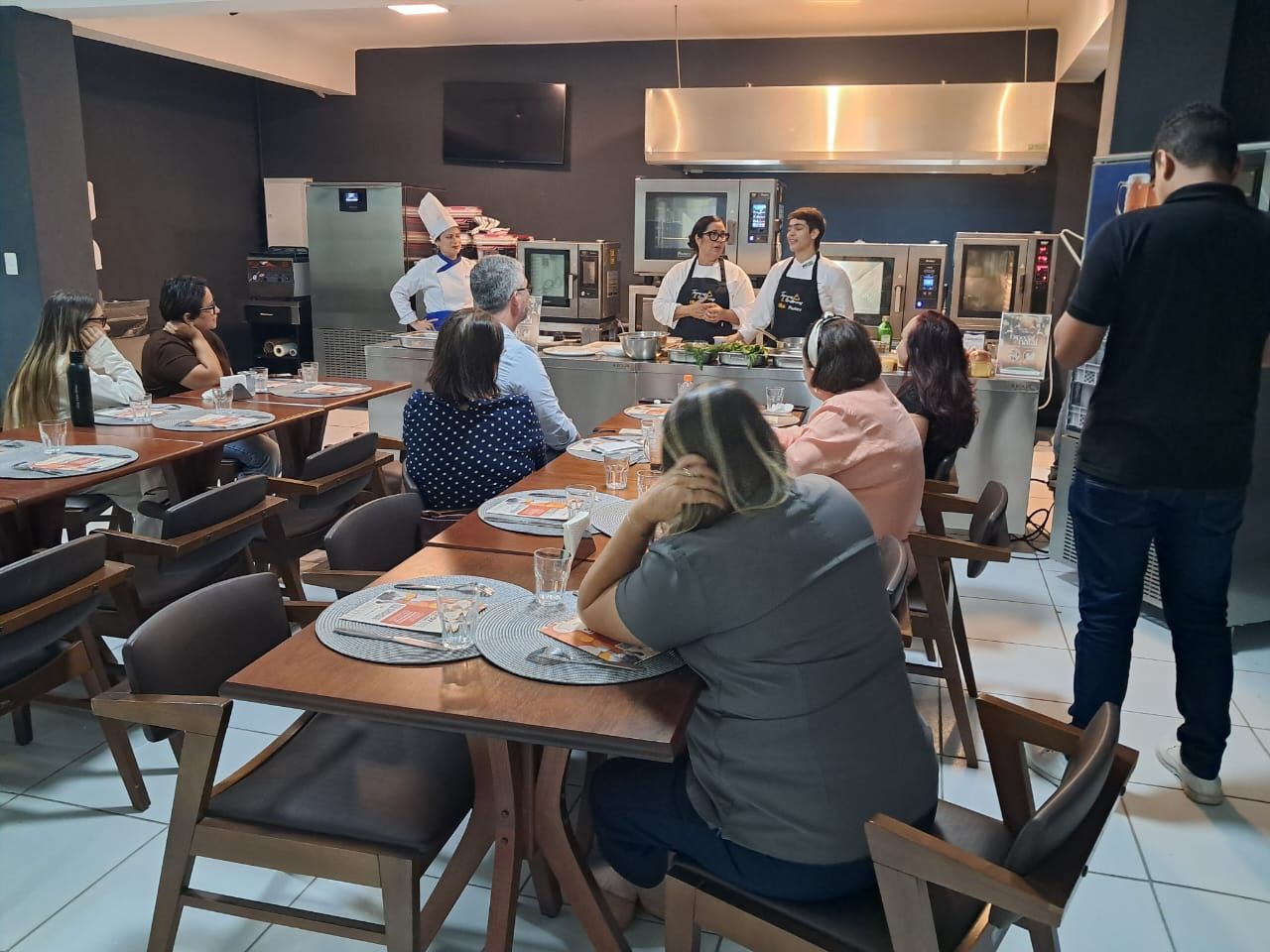 Aula de culinária: chefs demonstram pratos. Participantes observam nas mesas. Cozinha ao fundo.