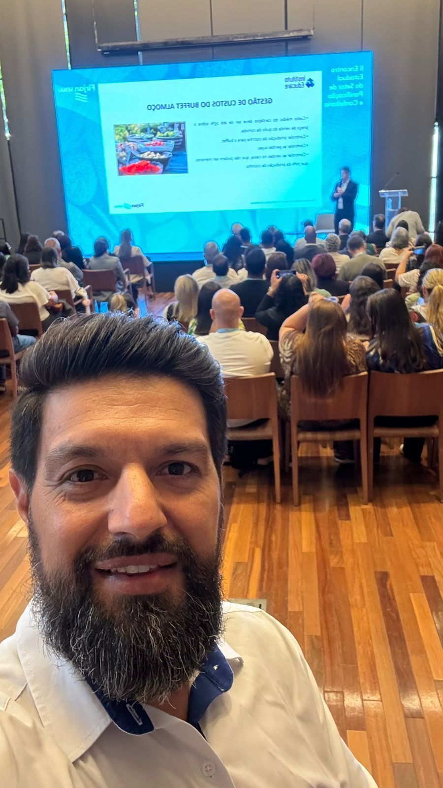 Homem tira selfie em uma conferência. Um palestrante faz uma apresentação em uma tela para um grande público.