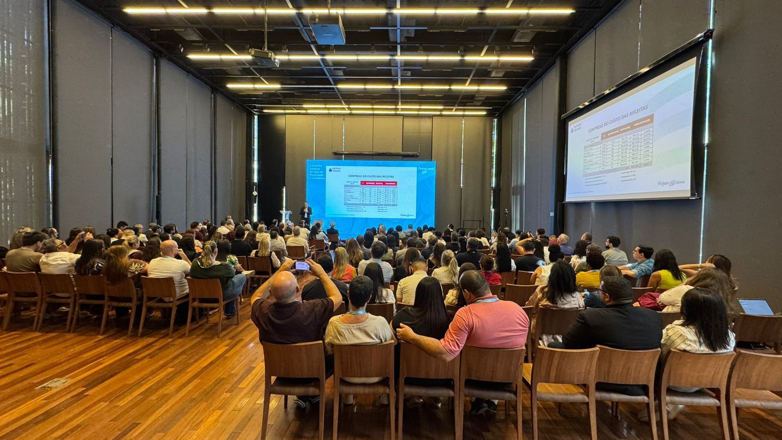 Público em uma conferência em uma sala grande, assistindo a uma apresentação em uma tela.