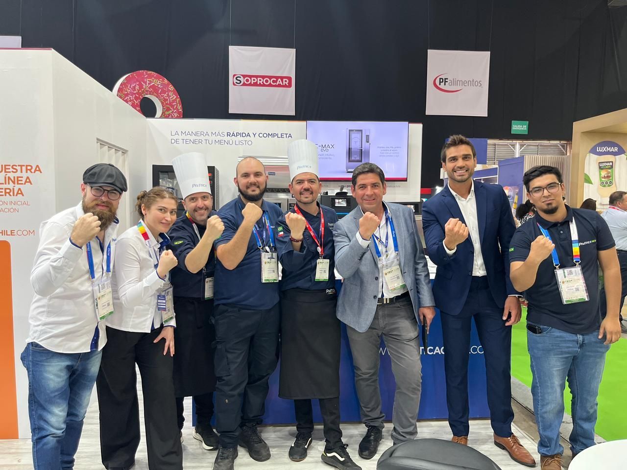 Grupo de nove pessoas comemorando em um estande de feira comercial, algumas usando chapéus de chef, sorrindo e flexionando os braços.