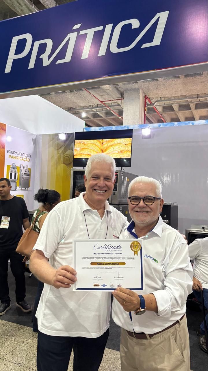 Dois homens sorrindo, segurando um certificado em um estande de feira comercial, placa