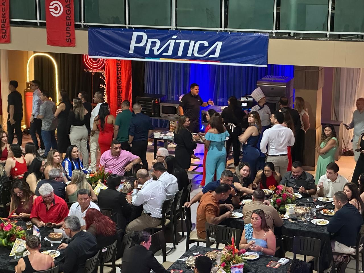 Pessoas em um evento formal, comendo em mesas, sob uma faixa que diz