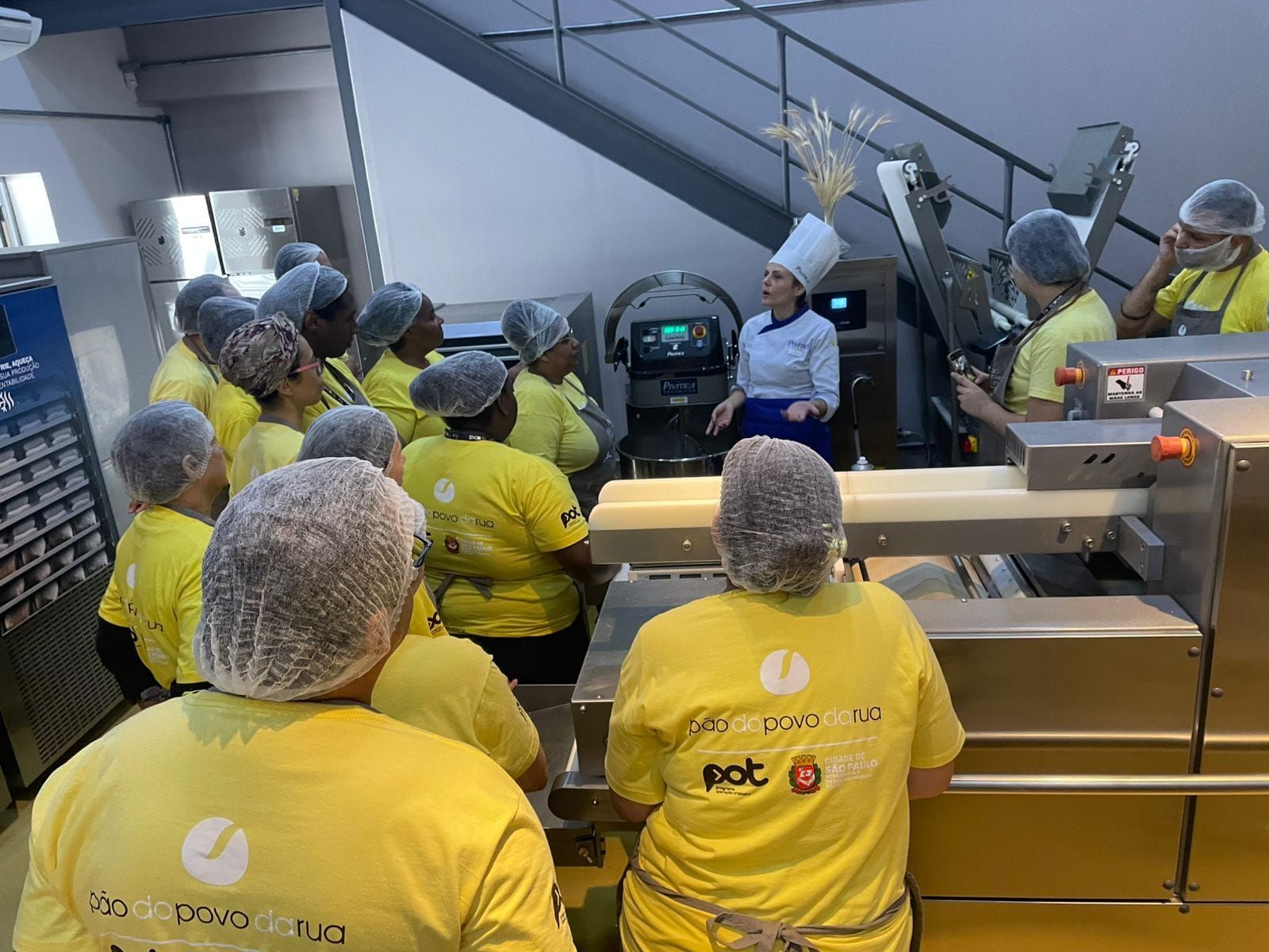 Um grupo de pessoas com redes de cabelo e camisas amarelas observa um chef demonstrando uma máquina em uma cozinha comercial.