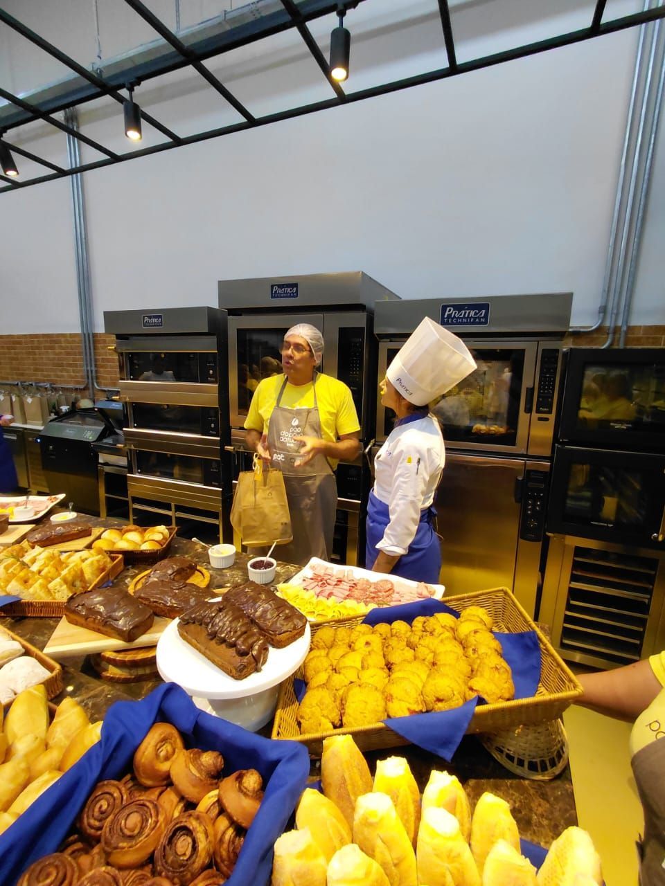 Uma exposição de produtos de padaria, duas pessoas conversando em frente aos fornos.