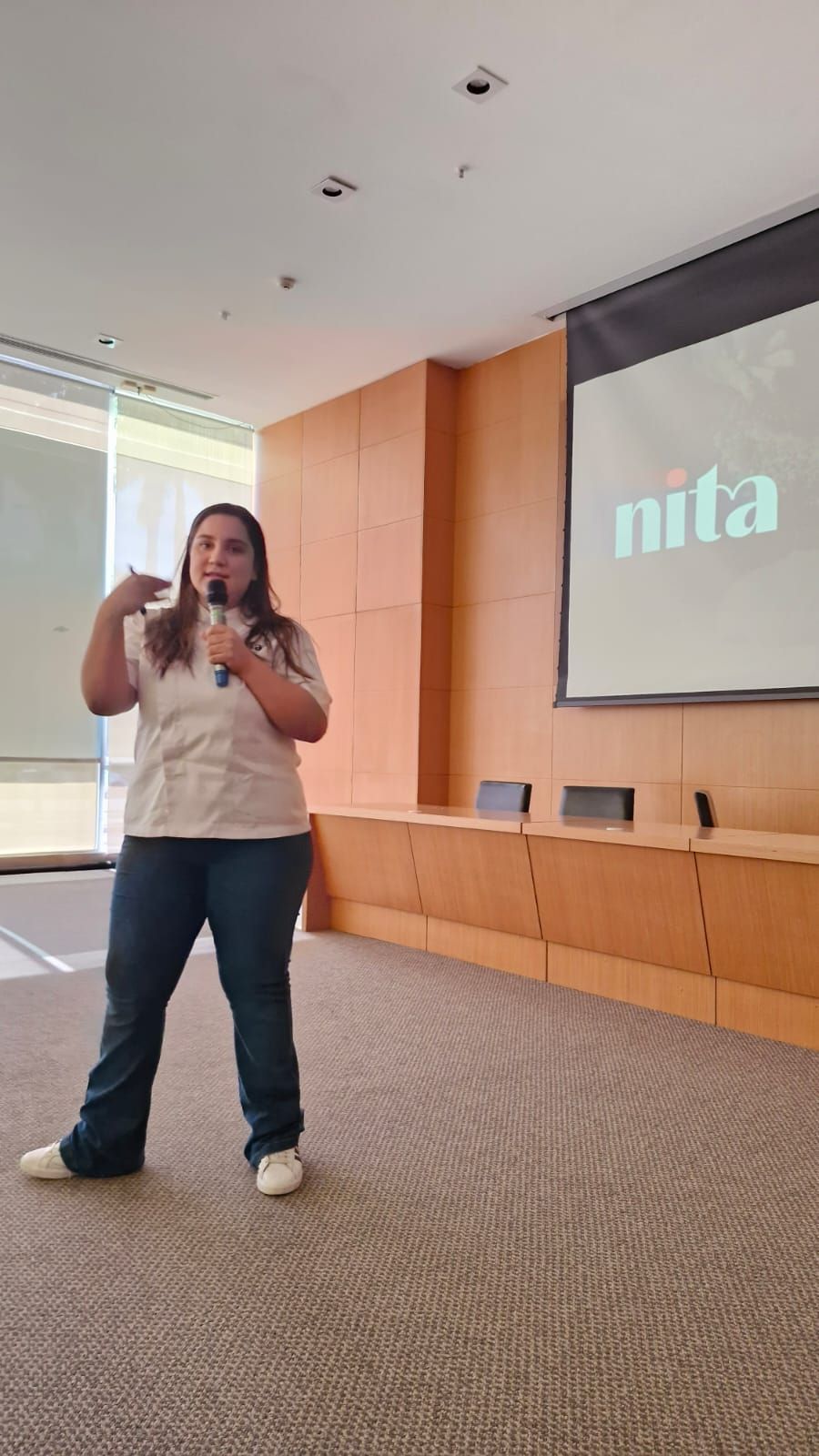 Mulher falando em uma apresentação, gesticulando, com o logotipo 