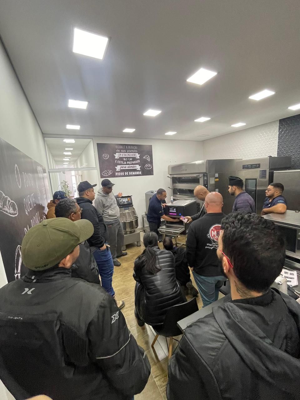Pessoas observando uma demonstração em uma cozinha comercial; uma apresentação está sendo feita.