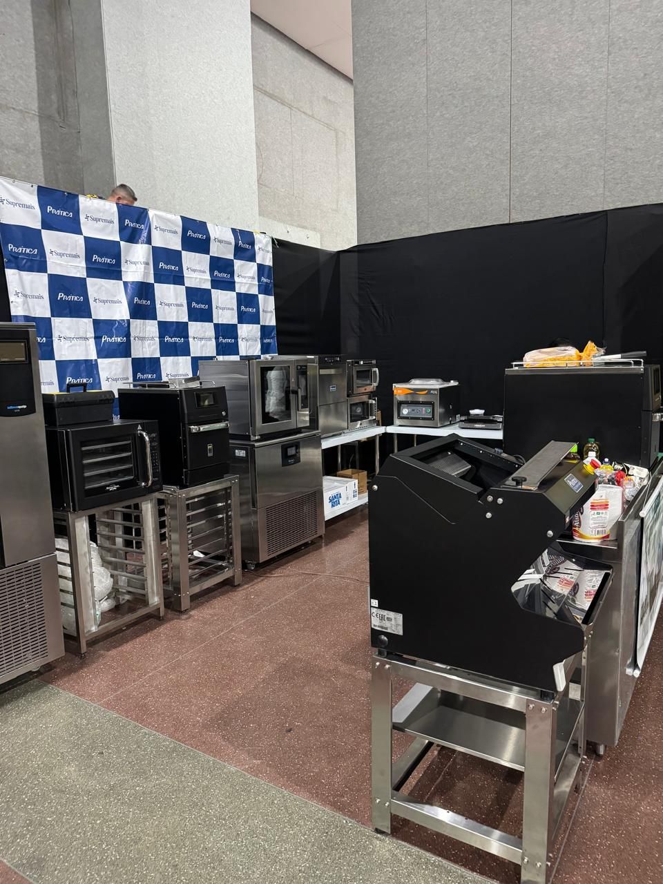 Um estande de feira comercial exibe equipamentos de cozinha comercial; máquinas de aço inoxidável e pretas com um fundo xadrez azul.