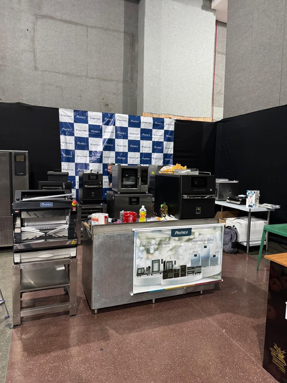 Exposição de equipamentos de cozinha comercial em um evento, eletrodomésticos de aço inoxidável em um balcão com um banner xadrez ao fundo.