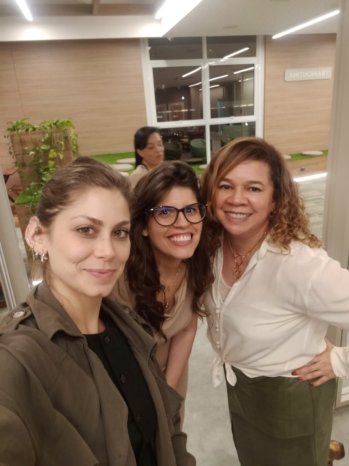 Três mulheres sorrindo para uma selfie em um ambiente interno, perto de uma grande janela. Uma delas está ao fundo.