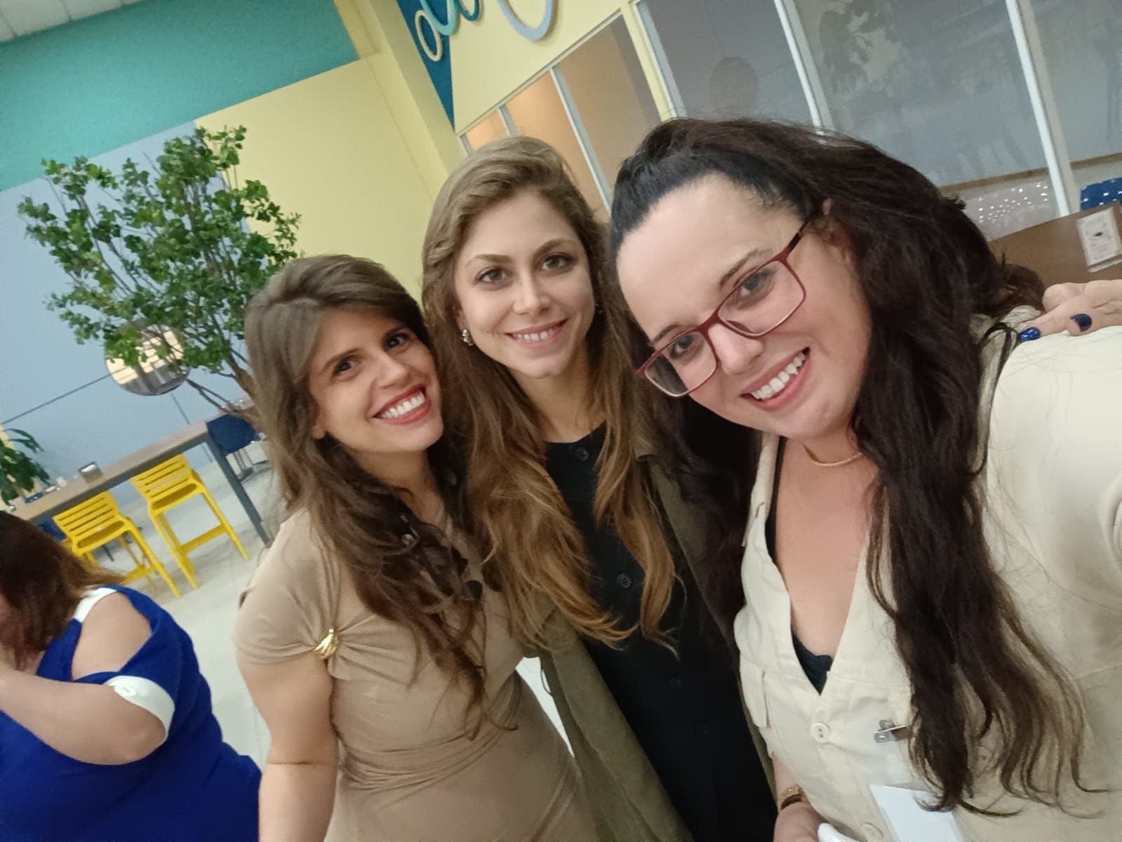 Três mulheres sorridentes tiram uma selfie dentro de casa perto de uma planta. Uma delas usa óculos e uma blusa bege.