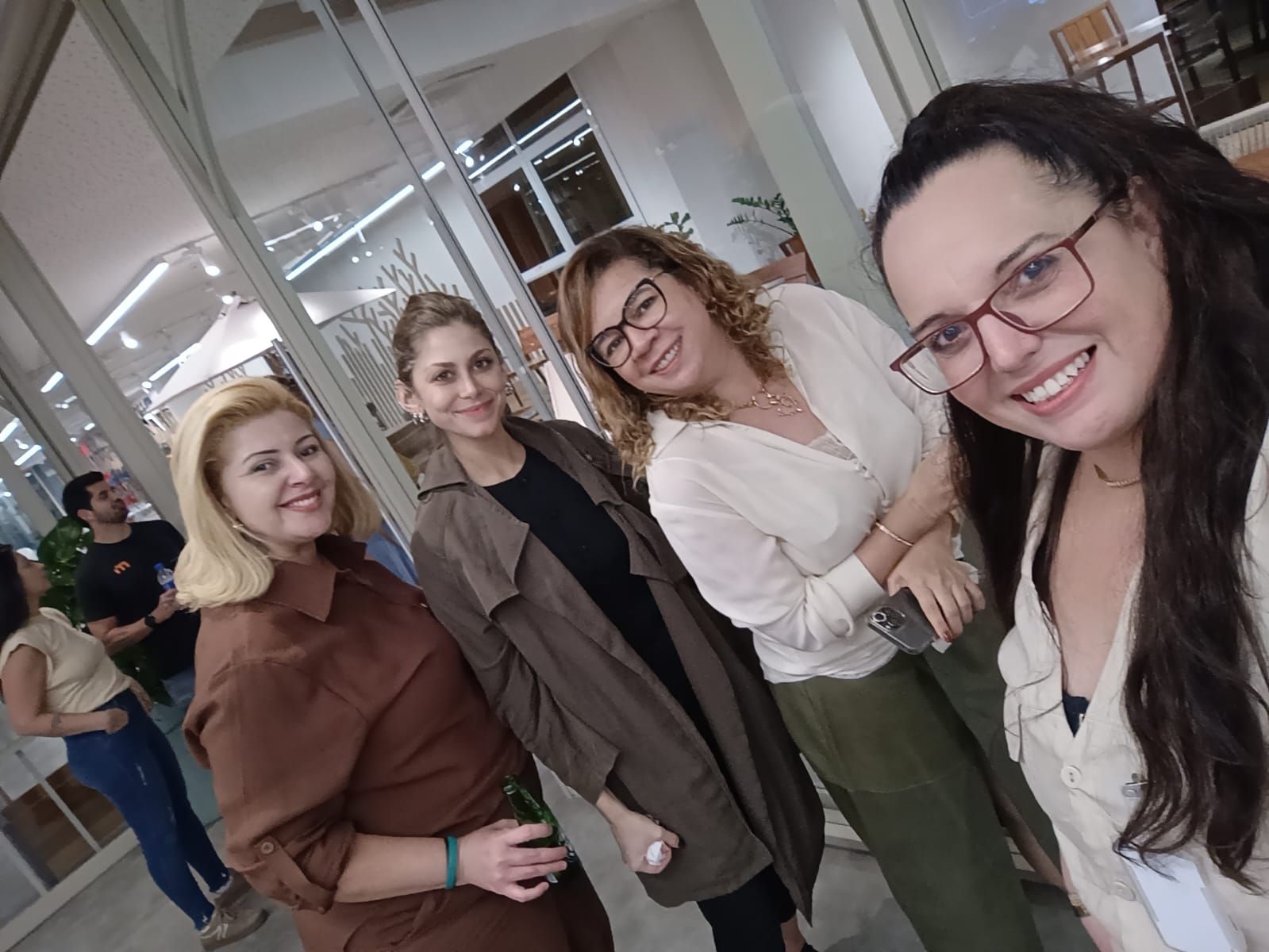 Quatro pessoas sorridentes posando juntas em um ambiente interno; três mulheres à direita e uma mulher à esquerda, vestindo cores variadas: bege, marrom e verde.
