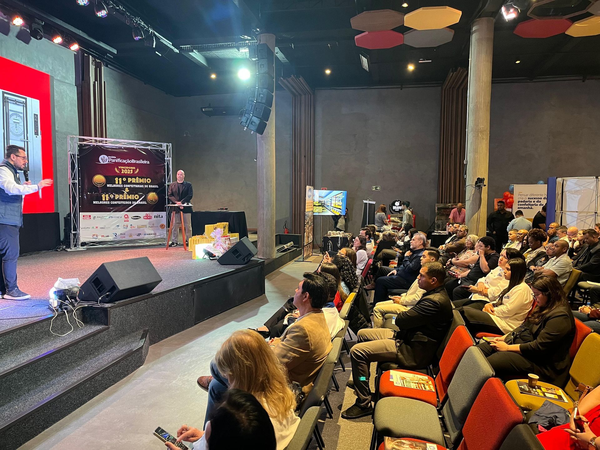 Um orador gesticula no palco durante uma conferência, dirigindo-se a uma plateia sentada em um grande auditório.