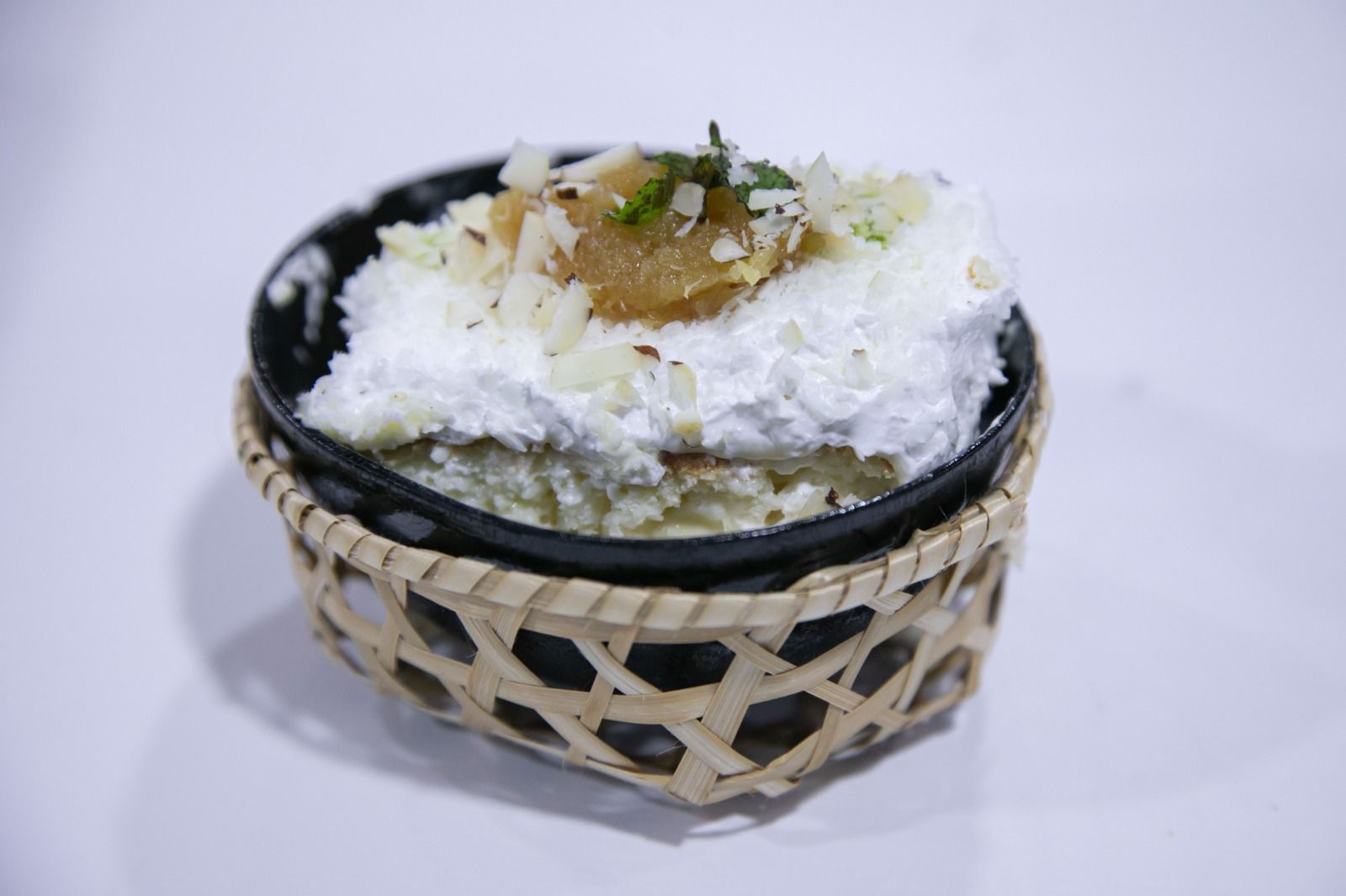 Sobremesa em uma cesta de vime; cobertura de creme branco, nozes, recheio marrom, tigela preta.