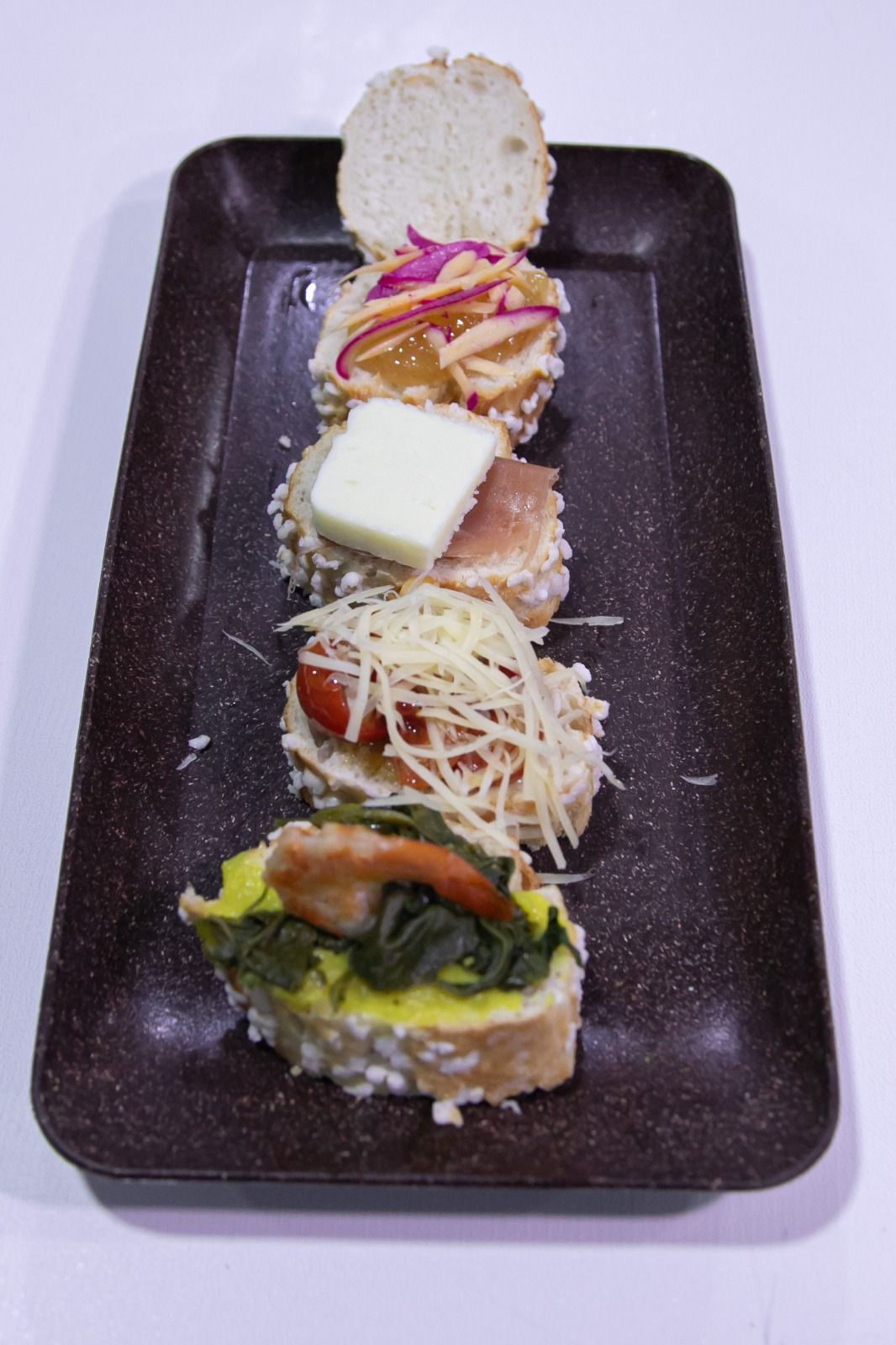 Cinco mini sanduíches abertos em um prato retangular marrom, cada um com recheios diferentes.