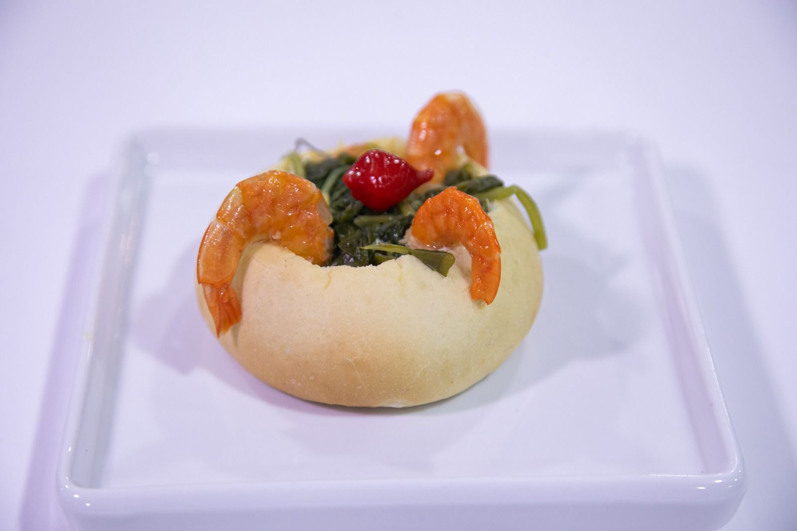 Prato de camarão e espinafre servido em uma tigela de pão, decorado com uma cereja, em um prato branco.