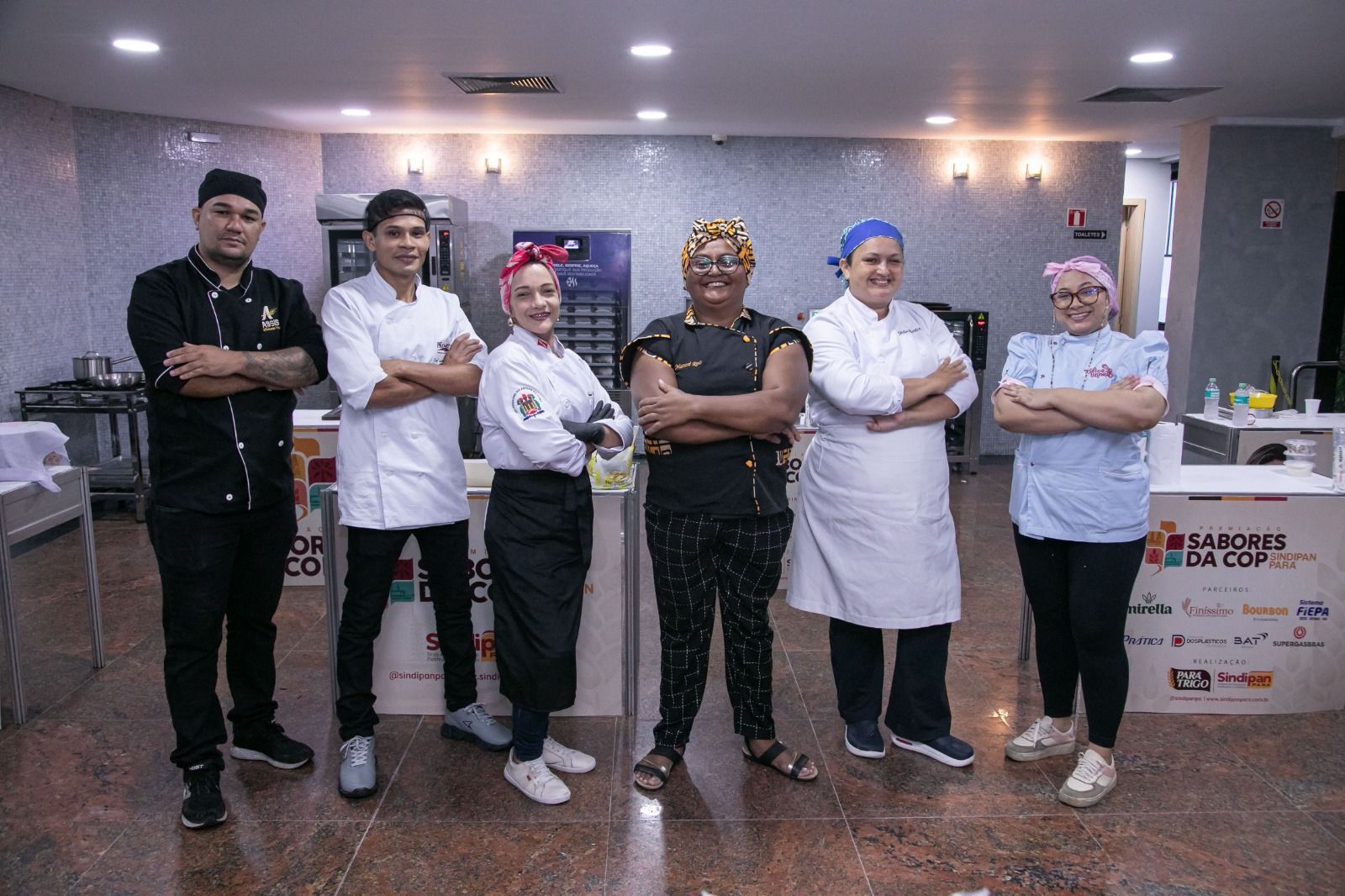 Seis chefs em uniformes variados posam com confiança em um cenário de cozinha, de braços cruzados.
