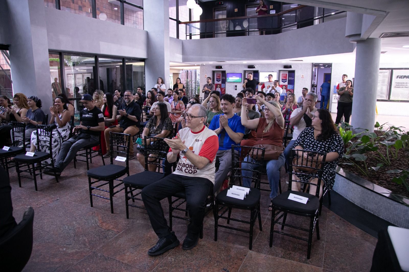 Pessoas aplaudindo em um evento em local fechado com assentos, plantas e vista do segundo andar.