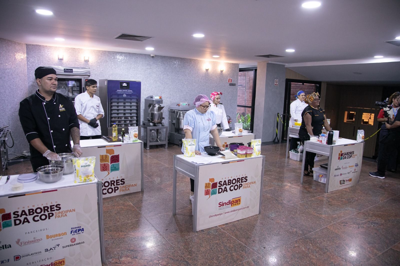 Chefs em estações de cozinha em uma sala bem iluminada para uma competição culinária.