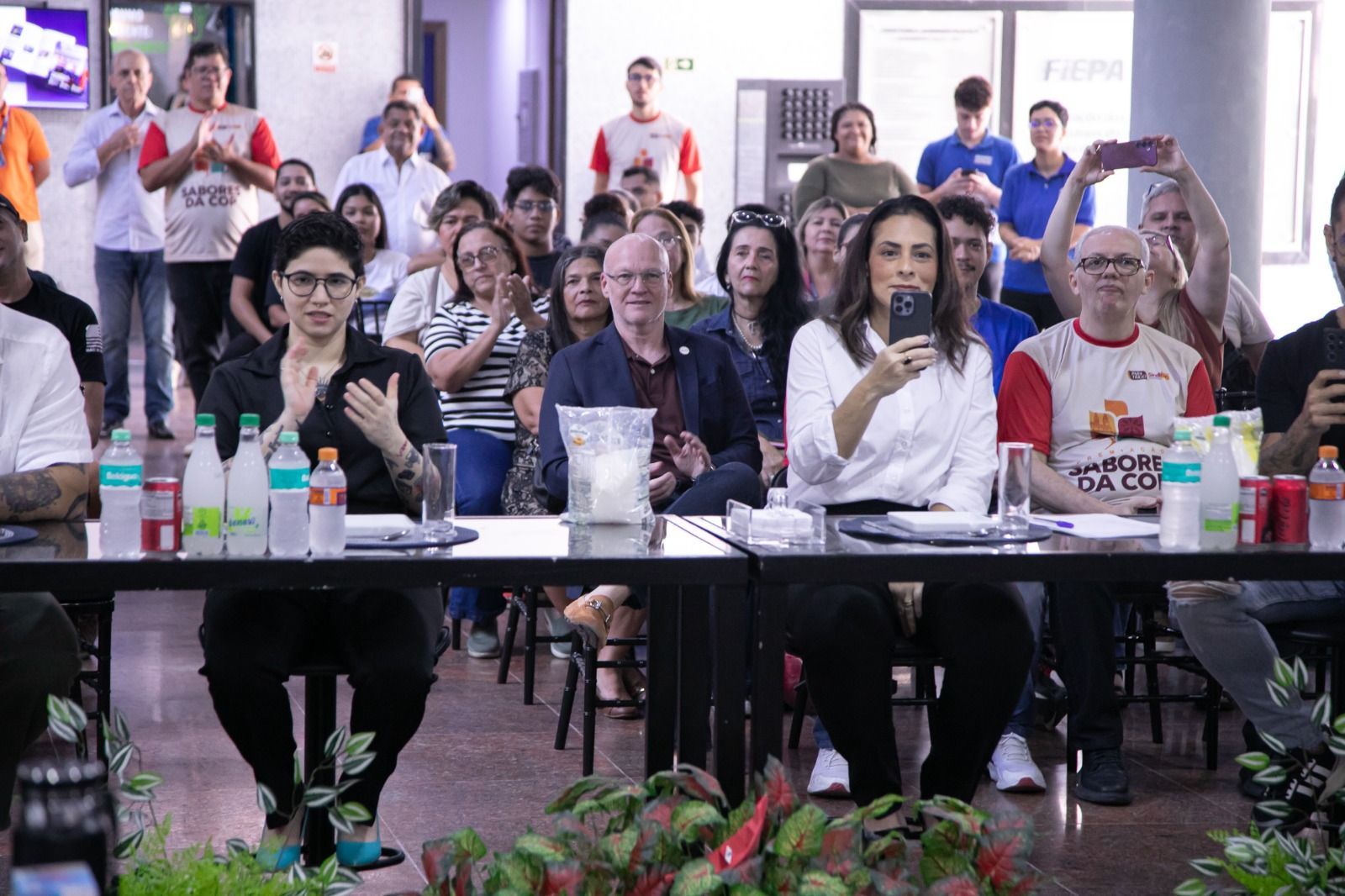 Pessoas sentadas à mesa aplaudem, duas mulheres e um homem no centro. Outras pessoas ficam de pé e aplaudem ao fundo.