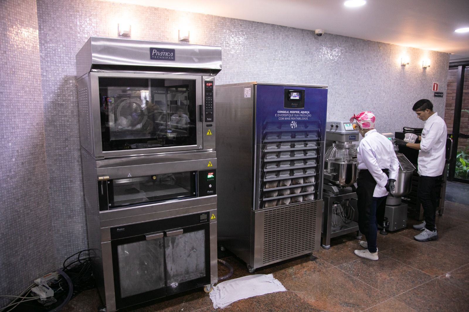 Cozinha com chefs perto de fornos industriais e um freezer, preparando alimentos.