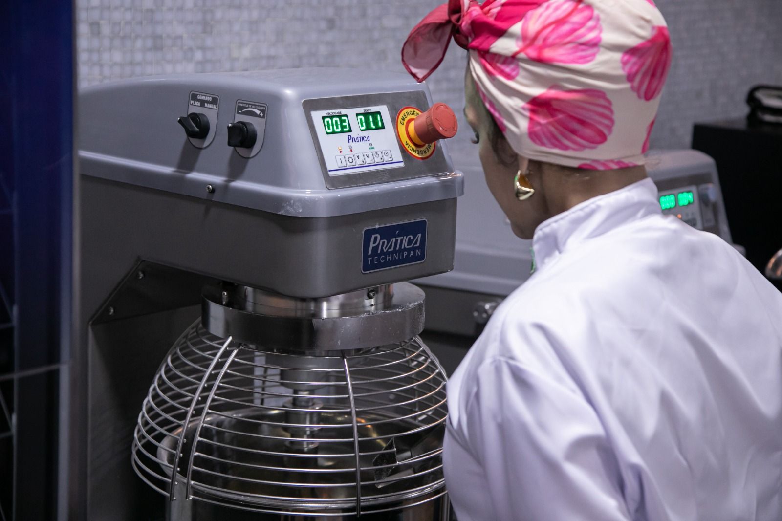 Mulher de jaleco branco e lenço rosa na cabeça operando uma batedeira industrial em uma cozinha.