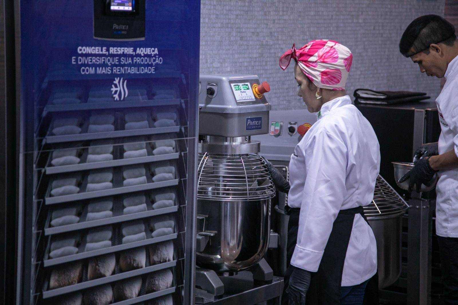 Duas pessoas em uma padaria, perto de uma grande batedeira e de um depósito com prateleiras de produtos embalados.