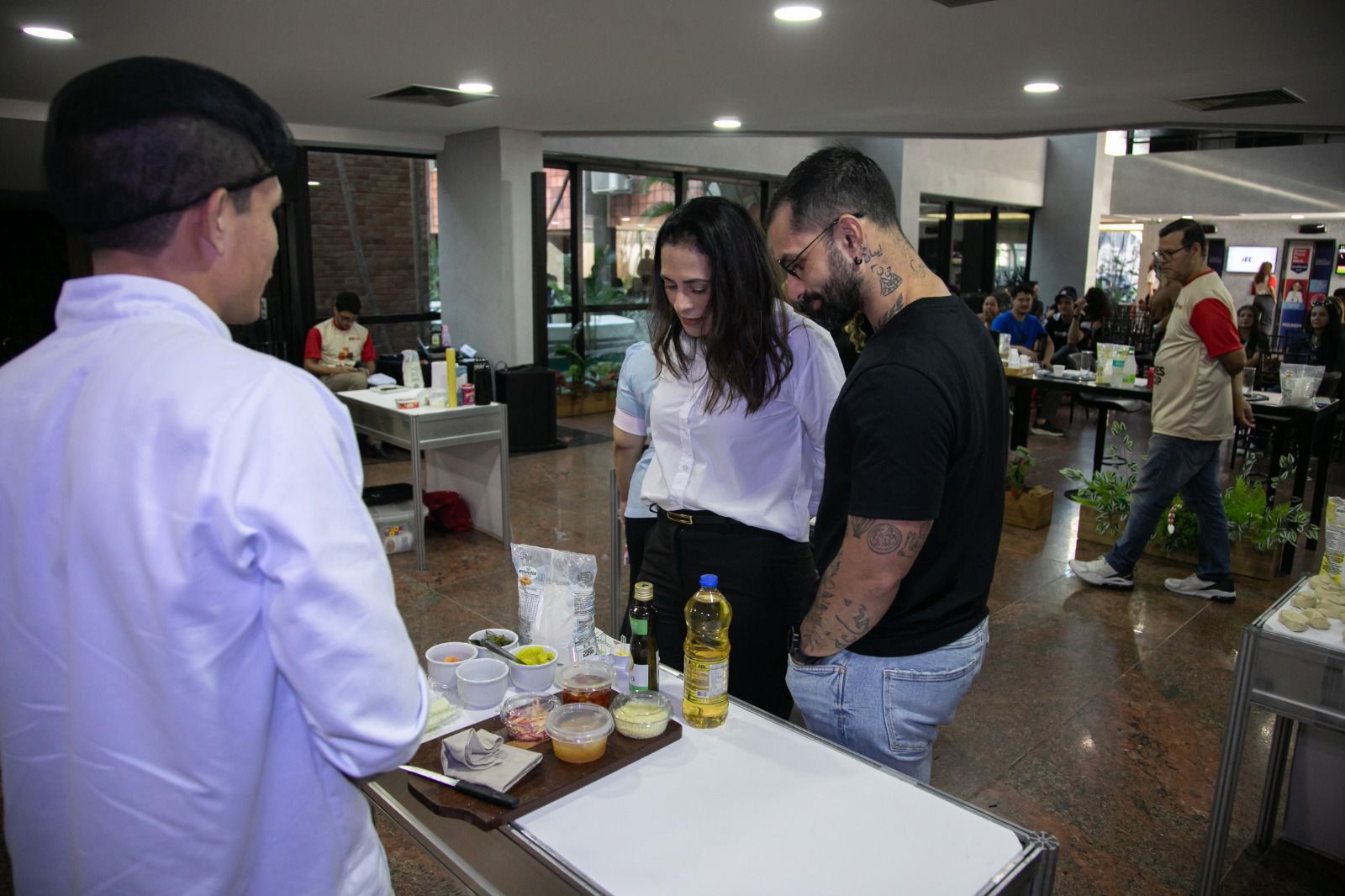 Chef demonstra técnicas culinárias para duas pessoas em uma cozinha comercial, com ingredientes e outros participantes.