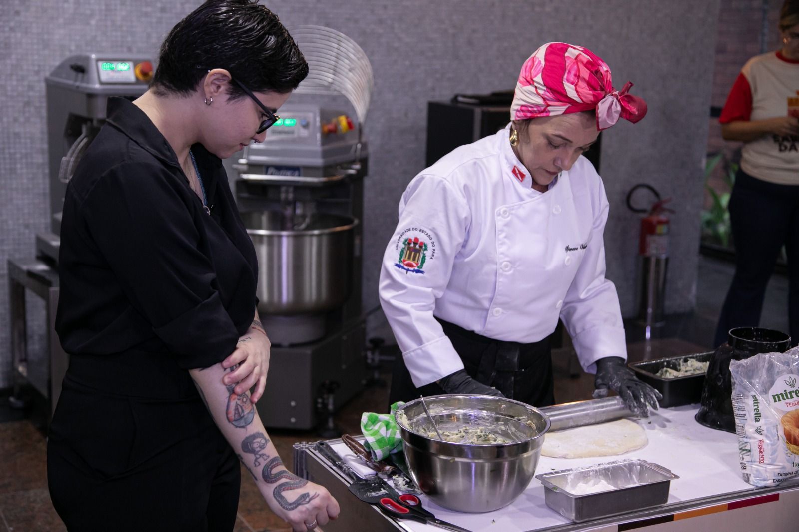 Em uma padaria, um chef de jaleco branco estende a massa no balcão enquanto outra pessoa observa.