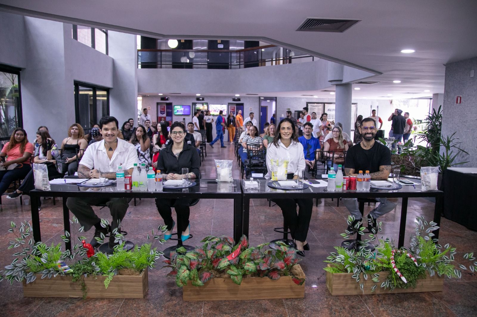 Painel com pessoas sentadas à mesa, plantas à frente e público atrás. Interior de um edifício.