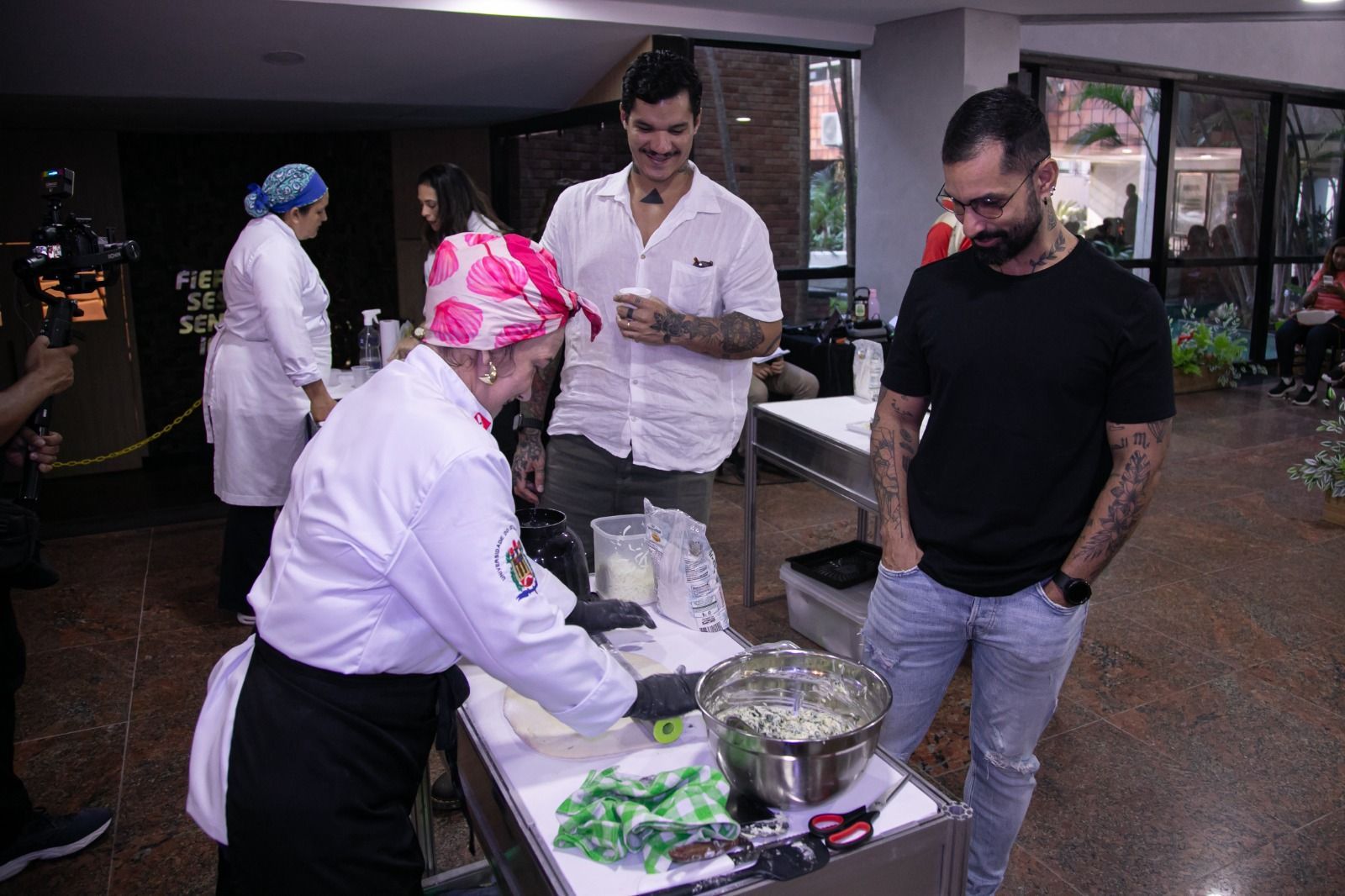 Chef preparando comida enquanto dois homens observam. Ambiente interno, pessoas ao fundo.