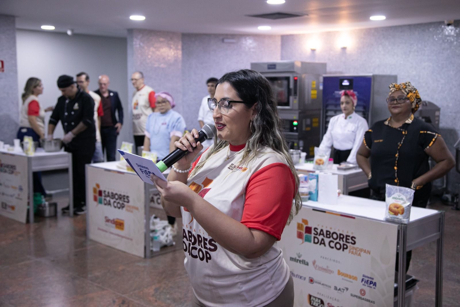 Mulher falando ao microfone em uma competição culinária, com chefs e preparo de alimentos ao fundo.
