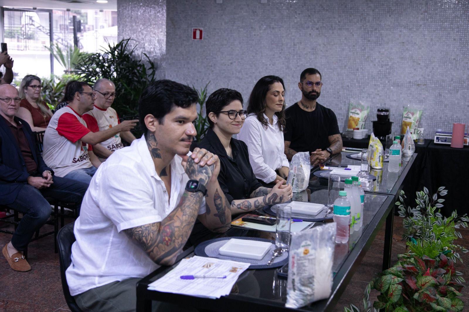Pessoas sentadas à mesa, algumas com tatuagens, parecem estar avaliando produtos. Copos e garrafas de água estão visíveis.