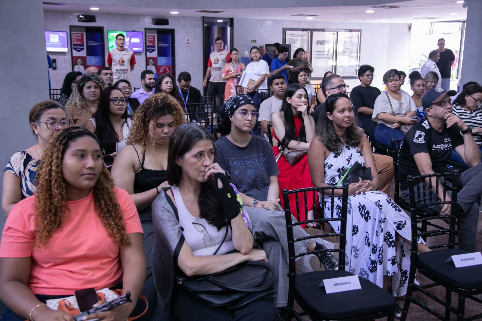 Público sentado, ouvindo atentamente. Ambiente interno, espaço moderno. Pessoas com estilos de vestimenta variados, expressões concentradas.