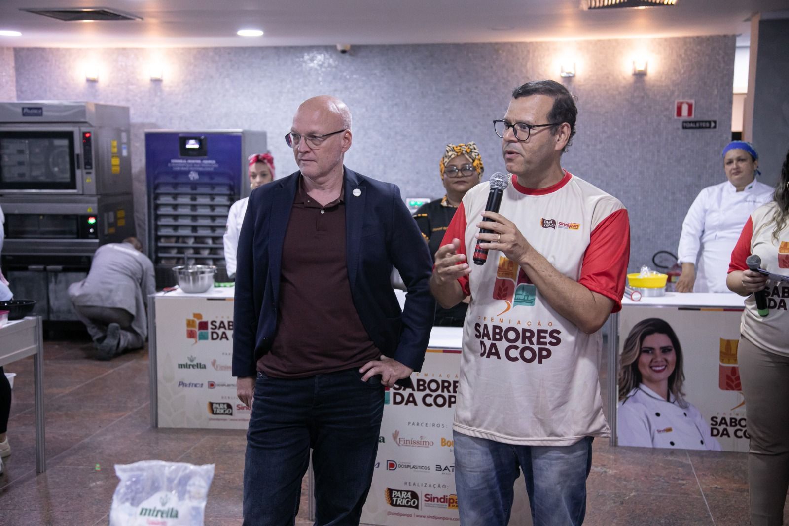 Dois homens conversando em um evento realizado em uma cozinha; um deles segura um microfone.