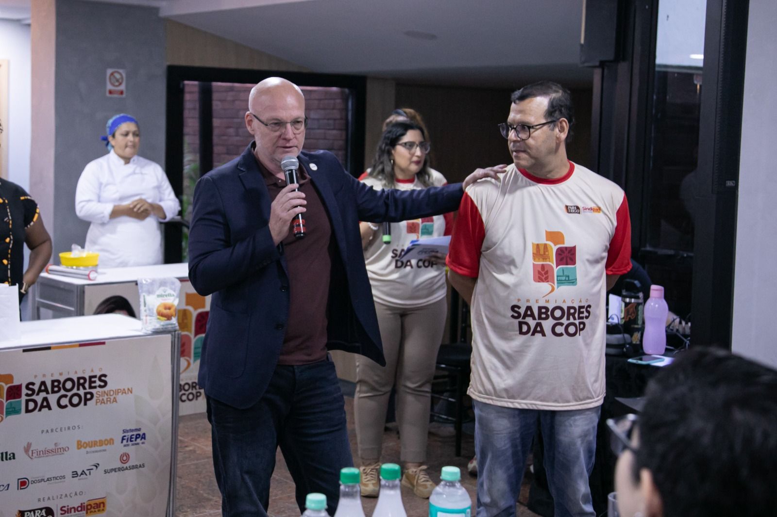 Um homem fala ao microfone e dá um tapinha no ombro de outro. Outros vestem camisetas com a marca da empresa. Evento em local fechado.