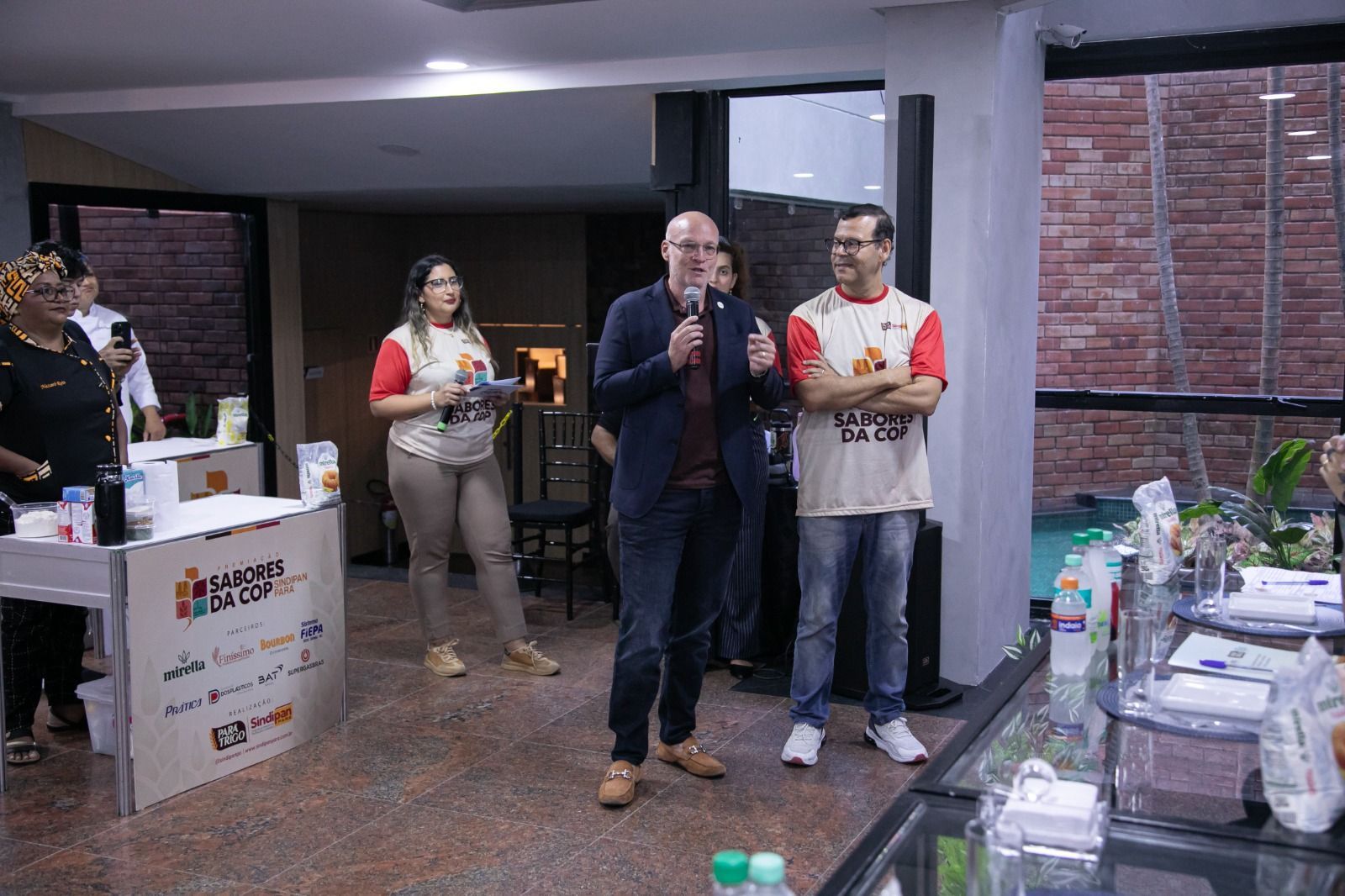 Homem discursando em evento, ladeado por duas pessoas, outras ao fundo. Sala com exposições.