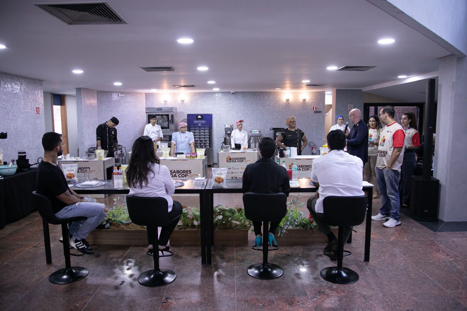Pessoas em uma competição culinária. Os jurados estão sentados às mesas observando os participantes, com estações de culinária em uma sala bem iluminada.