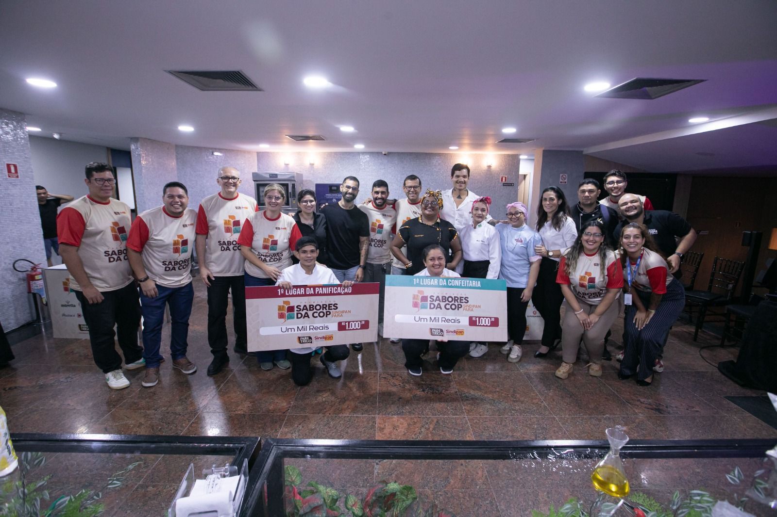 Grupo de pessoas segurando cartazes em ambiente fechado, possivelmente após um evento. Os cartazes contêm texto e logotipos.