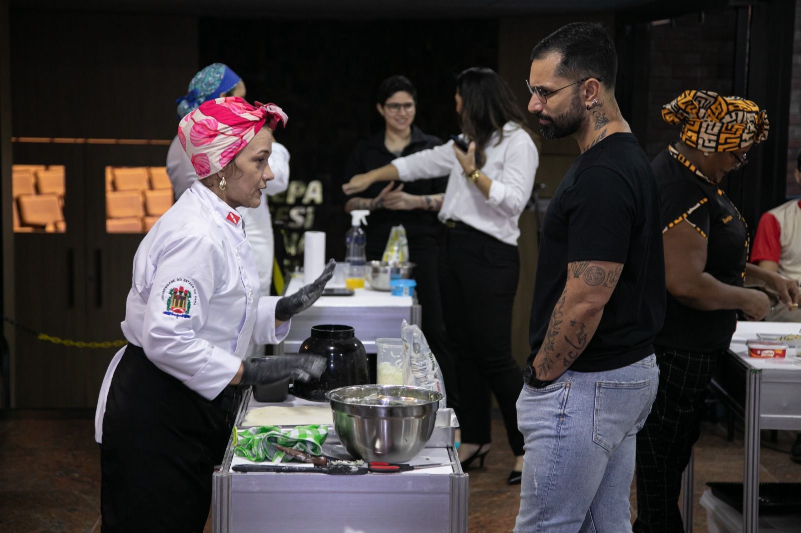 Um chef de jaleco branco discute um prato com um homem de camisa preta; outras pessoas assistem a uma competição culinária.