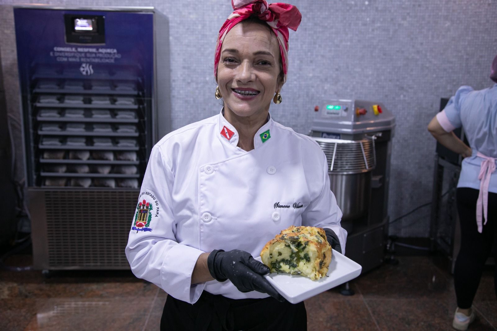 Um chef de cozinha, de jaleco branco e lenço na cabeça, segura um prato de comida, sorrindo, em meio à cozinha.