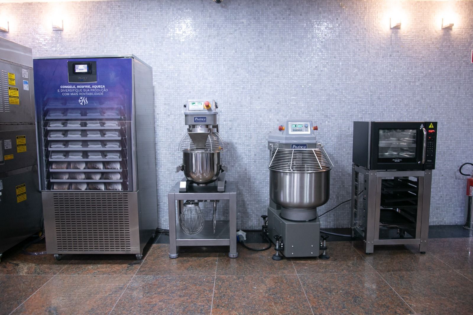 Conjunto de eletrodomésticos de cozinha comercial: freezer, batedeiras e forno encostados em uma parede revestida de azulejos.